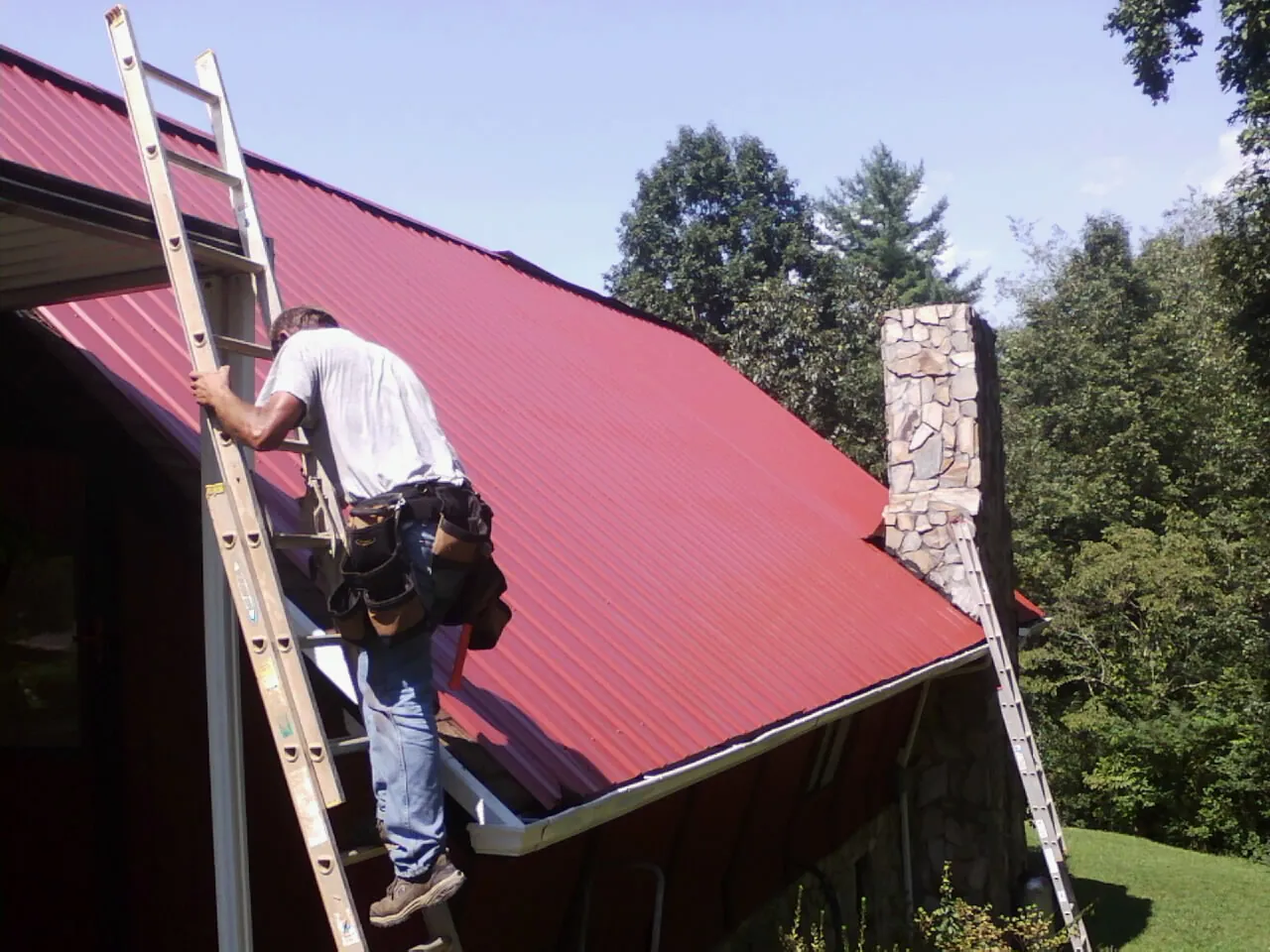 Roofer on a ladder beside a red metal roof and stone chimney — Soffit & Fascia Repair by Roofing Brothers in Pomaria